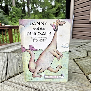 Puede incluir: Un libro infantil vintage titulado "DANNY and the DINOSAUR" de Syd Hoff. La portada presenta un dinosaurio de dibujos animados con un niño en su cuello. El libro es un "I CAN READ Book" con una ilustración colorida.