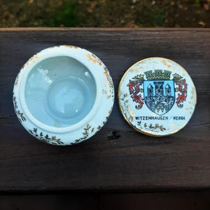May include: A small, open, white ceramic trinket box with a lid. The box and lid are decorated with gold floral patterns. The lid features a crest with a castle, and the text "WITZENHAUSEN / WERRA".