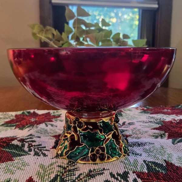 Pode incluir: Uma taça de vidro vermelho profundo apoiada numa base decorativa com acabamento dourado metálico, folhas de azevinho verdes e bagas vermelho-escuras. A taça tem uma superfície lisa e translúcida e uma abertura circular larga.
