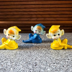 Puede incluir: Tres figuras de elfos de cerámica. Dos son amarillos, uno es azul. Tienen orejas blancas, mejillas rosadas y pelo naranja y blanco. Los elfos están sentados, con las piernas extendidas.