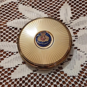 May include: A gold compact mirror with a blue and red enamel crest featuring a crown and the text "Queen Elizabeth 2".