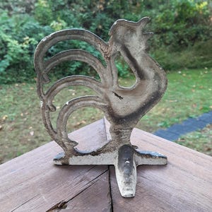 May include: A white cast iron rooster doorstop with a detailed design. The rooster is standing on a three-pronged base.