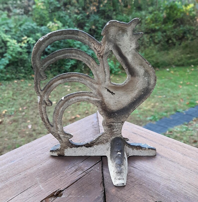 May include: A white cast iron rooster doorstop with a detailed design. The rooster is standing on a three-pronged base.