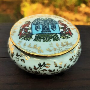 May include: A small, round porcelain trinket box with a lid. The box is white with gold trim and floral designs. The lid features a blue and gold crest design with red accents. Likely vintage or antique.