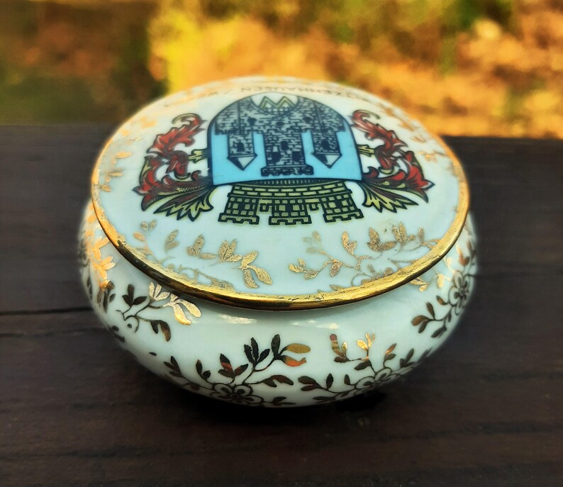 May include: A small, round porcelain trinket box with a lid. The box is white with gold trim and floral designs. The lid features a blue and gold crest design with red accents. Likely vintage or antique.