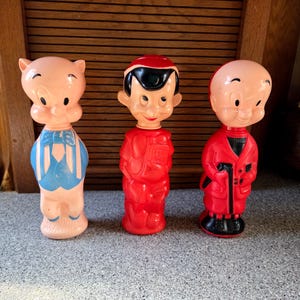 Puede incluir: Tres figuras de personajes de dibujos animados vintage. El primero es un cerdo rosa con un chaleco azul con rayas blancas. El segundo es un niño vestido de rojo con una gorra negra. El tercero es una figura vestida de rojo con zapatos negros.