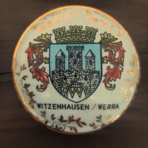 May include: A circular, white ceramic decorative plate with a gold rim and floral detailing. The centre displays a coat of arms with a castle design in blue, black, and white, and the text "WITZENHAUSEN / WERRA".