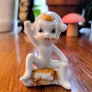 May include: A small, white ceramic figurine of a seated figure with a raised arm. The figure has a small orange patch on its head and is seated on a brown and white base. The figurine has black eyelashes and a red mouth.