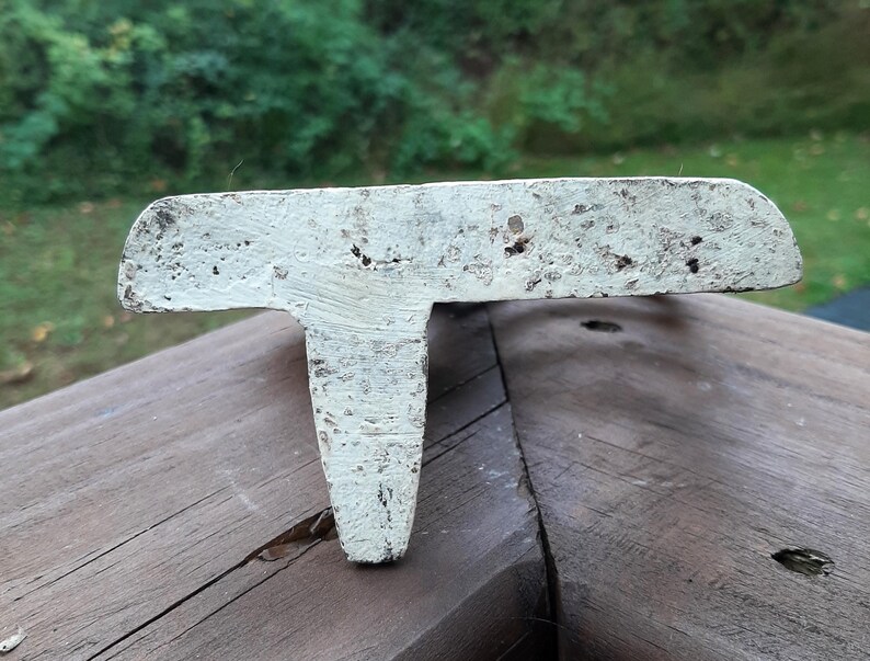May include: White cast iron doorstop shaped like a T. The doorstop has a distressed finish and is sitting on a wooden surface.