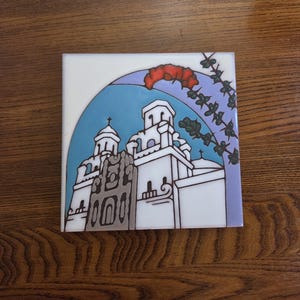 May include: Square ceramic tile featuring a white church with two bell towers, a blue sky, and a red floral accent. The church has black outlines and gray details. A purple and green floral design frames the top right corner.