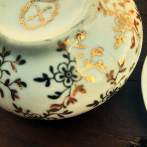 May include: Close-up of a white porcelain teacup with gold floral and leaf designs. The cup features a gold emblem near the rim. The floral pattern includes black and red accents. The cup is on a dark brown surface.