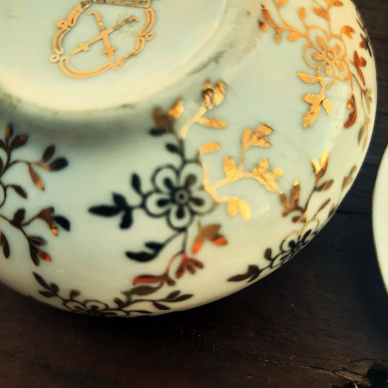 May include: Close-up of a white porcelain teacup with gold floral and leaf designs. The cup features a gold emblem near the rim. The floral pattern includes black and red accents. The cup is on a dark brown surface.
