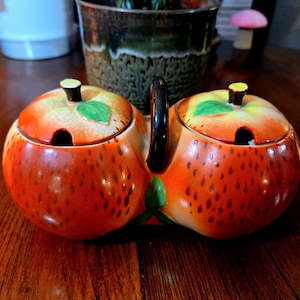 May include: A ceramic apple-shaped double condiment server with a black handle. The server is orange with brown speckles, green leaf accents, and yellow and green lids. Each lid has a small black knob and a spoon opening.