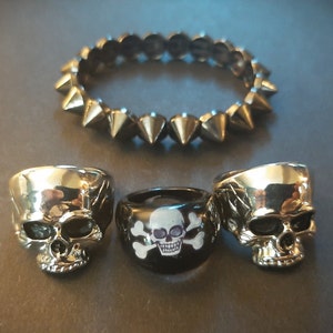 May include: Three silver skull rings, one with a black skull and crossbones design, and a silver bracelet with spikes.