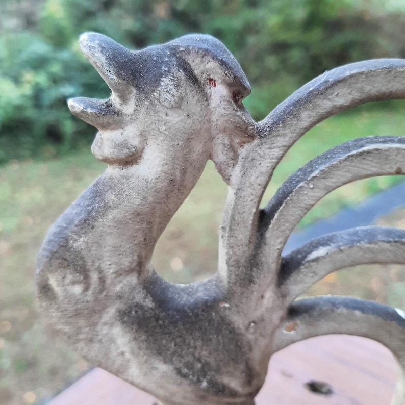 May include: A weathered gray cast iron rooster figurine with a raised tail and a detailed head. The rooster is facing to the right and has its beak open.