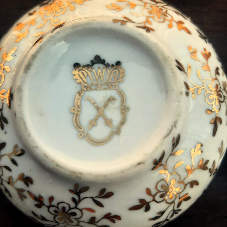 May include: A white porcelain saucer with gold floral designs and a gold emblem. The emblem features a crown above the letters "PWB" and crossed swords. The saucer's rim is decorated with gold floral patterns.