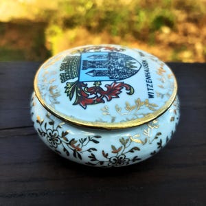 May include: A small, round, white ceramic trinket box with a lid. The lid features a crest with a blue and white design and the text "WITZENHAUSEN". Gold accents and floral patterns adorn the box.