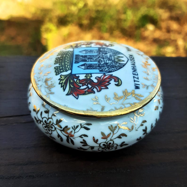 May include: A small, round, white ceramic trinket box with a lid. The lid features a crest with a blue and white design and the text "WITZENHAUSEN". Gold accents and floral patterns adorn the box.