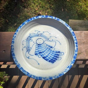 Puede incluir: Un plato redondo de cerámica con un diseño de ángel azul y blanco. El ángel tiene un chal a cuadros y está rodeado por una corona. El borde del plato tiene un borde azul moteado. El plato está sobre una superficie de madera.