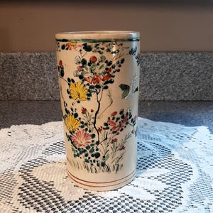 May include: A white ceramic vase with a floral design. The vase has a cylindrical shape and is decorated with green, yellow, red, and blue flowers and birds.