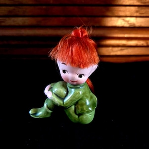 Puede incluir: Figura de cerámica de un elfo sentado con el pelo rojo brillante recogido en un moño. El elfo lleva un traje verde y tiene una cara sonriente con rasgos delicados. El fondo es una superficie oscura con un telón de fondo de listones de madera.