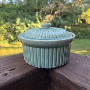 Könnte beinhalten: Eine türkisfarbene Keramik-Auflaufform mit Deckel. Die Schüssel hat ein geripptes Design an den Seiten und am Deckel sowie einen kleinen Griff oben. Die Schüssel steht auf einer Holzoberfläche.