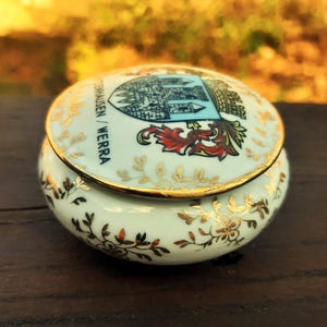 May include: A small, round, ceramic trinket box with a lid. The box is light blue with gold floral accents and a gold rim. The lid features a crest with the text "TENHAUSEN / WERRA".
