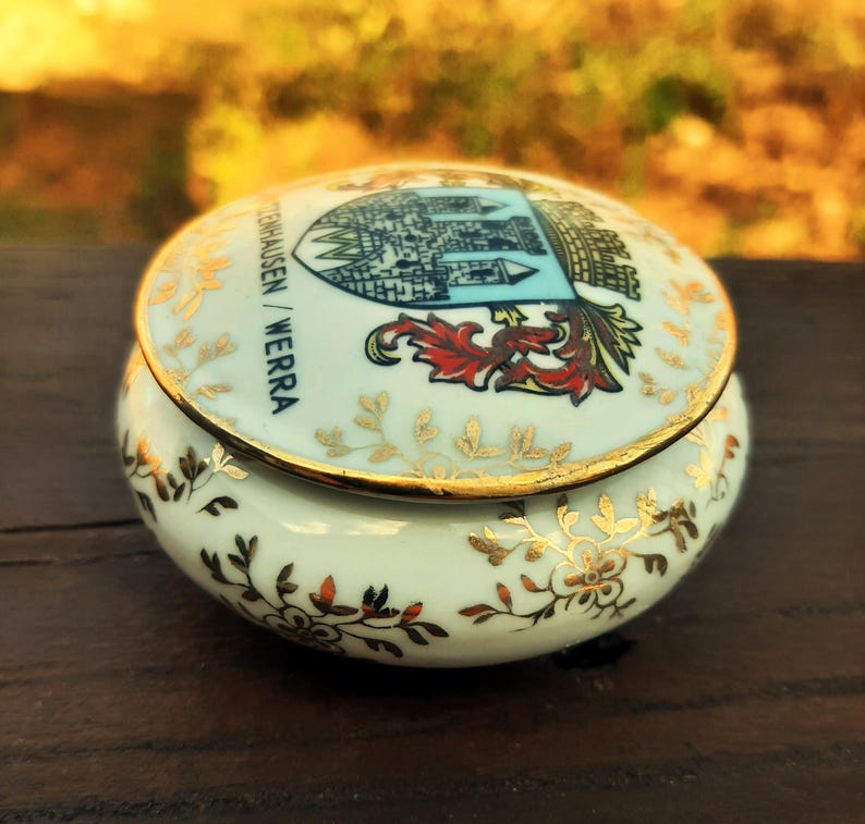May include: A small, round, ceramic trinket box with a lid. The box is light blue with gold floral accents and a gold rim. The lid features a crest with the text "TENHAUSEN / WERRA".