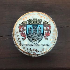 May include: A circular, white ceramic trinket box with a gold rim and floral detailing. The lid displays a coat of arms with a castle design and the text "WITZENHAUSEN / WERRA".