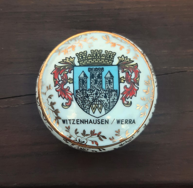 May include: A circular, white ceramic trinket box with a gold rim and floral detailing. The lid displays a coat of arms with a castle design and the text "WITZENHAUSEN / WERRA".