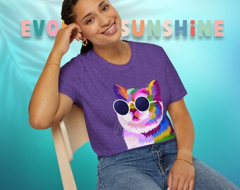 Op de afbeelding: Een paars t-shirt met korte mouwen en een afbeelding van een veelkleurige kat met een ronde zonnebril. Het shirt heeft een ronde hals en wordt gecombineerd met een blauwe spijkerbroek. De achtergrond toont de tekst "EVOLVE SUNSHINE" in pastel.
