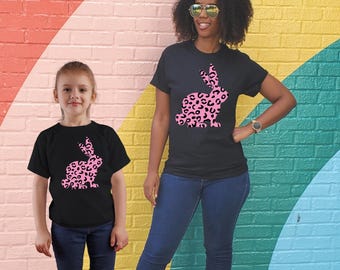 May include: Two people wearing black t-shirts with a pink leopard print bunny graphic. The bunny is facing the right side of the image. The person on the left is a child and the person on the right is an adult.