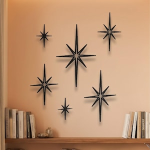 Könnte beinhalten: Ein Set aus sechs schwarzen Sternen-Wanddekorationen in verschiedenen Größen, die jeweils einen Mittelpunkt mit strahlenden Spitzen aufweisen. Diese geometrischen Ornamente sind in einer Gruppe an einer Wand angeordnet und zeigen ein symmetrisches Design.