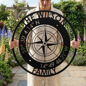 May include: Black metal family compass rose wall art. The design includes the text "THE WILSON FAMILY" and coordinates: 59° 30' 4.14" N and 14° 49' 46.21" W. The compass rose has a star design in the centre.