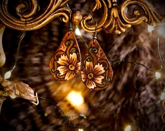May include: Teardrop-shaped leather earrings, hand-painted with a floral design in brown, tan, and green. The earrings hang from gold-coloured metal hooks. The background is blurred, with warm lighting and a soft, textured appearance.