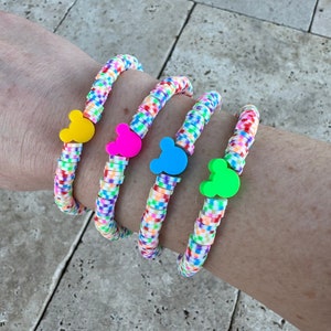 May include: Four colorful bracelets with Mickey Mouse head charms. The bracelets are made of small, colorful beads and are stacked on a person's wrist. The charms are in yellow, pink, blue, and green.