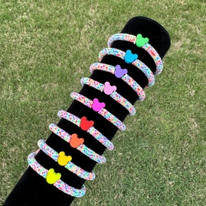 May include: A set of nine colorful bracelets with a Mickey Mouse head charm on each. The bracelets are made of a white string with colorful confetti-like dots. The charms are in different colors, including green, blue, purple, pink, red, orange, and yellow.