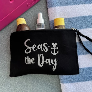 May include: Black canvas zippered pouch with a silver glitter design that says "Seas the Day" with an anchor and a Mickey Mouse head. The pouch has a black handle and is lying on a blue and white striped towel.