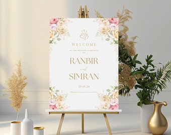 May include: A white wedding welcome sign with gold lettering and floral accents. The sign reads "Welcome to the wedding ceremony of Ranbir and Simran 29.05.24".