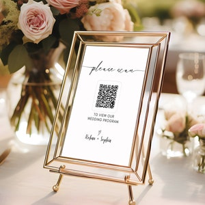 May include: A gold-framed sign with a white background and black text that reads "Please scan to view our wedding program" with a QR code and the names "Rebecca & Sophia".