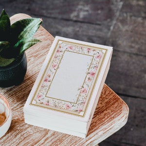 Puede incluir: Un libro blanco con un diseño floral en la portada. Las flores son rosas y blancas con detalles dorados. El libro está cerrado y se encuentra sobre una superficie de madera.