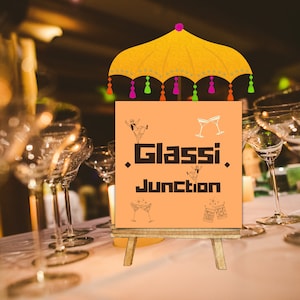 May include: A sign with the text "Glassie Junction" on a wooden easel with a yellow and orange umbrella above it. The sign is decorated with drawings of cocktails and dancing figures.