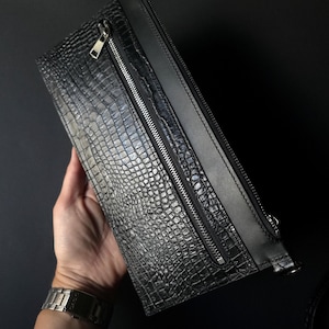 May include: Black clutch bag with a crocodile skin effect. The bag features a silver zip and a black leather strap. The rectangular bag appears to be made of leather.