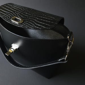 May include: Black crossbody bag with a textured flap and silver-tone hardware. The bag features a curved design and a long strap. The flap has a crocodile-like pattern. The interior is visible and has a striped lining.