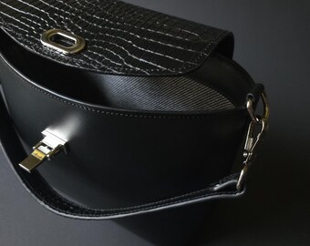 May include: Black crossbody bag with a textured flap and silver-tone hardware. The bag features a curved design and a long strap. The flap has a crocodile-like pattern. The interior is visible and has a striped lining.