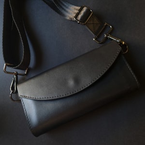 Könnte beinhalten: Eine schwarze Crossbody-Tasche aus Leder mit geschwungenem Klappenverschluss. Die Tasche hat einen verstellbaren schwarz-grau gestreiften Riemen mit silberfarbenen Metallbeschlägen. Die Tasche wird auf dunklem Hintergrund gezeigt.