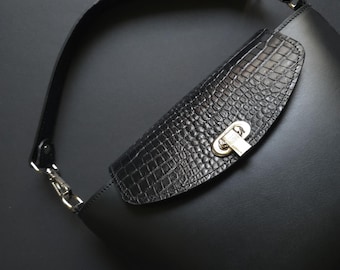 May include: Black leather handbag with a textured flap and silver-tone hardware. The bag features a long, adjustable strap and a secure clasp closure. The design combines smooth leather with a reptile-patterned accent.