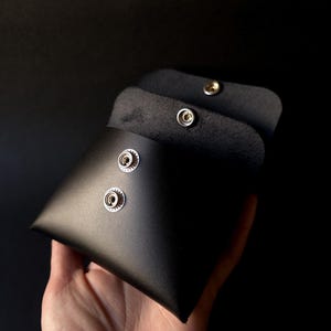 May include: A black leather pouch with a triangular shape, held open to reveal the interior. The pouch has two silver snap closures on the front and a third on the flap. The background is black.