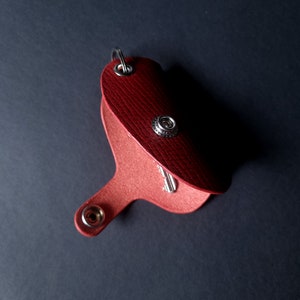 May include: A red leather key holder with a silver key ring and a silver snap closure. The key holder is open and a silver key is visible inside.