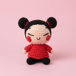 May include: A handmade crochet doll with a smiling face, black hair buns, and red accents on a pink background. The doll wears a red dress and black shoes. The doll is a cute, handcrafted item.